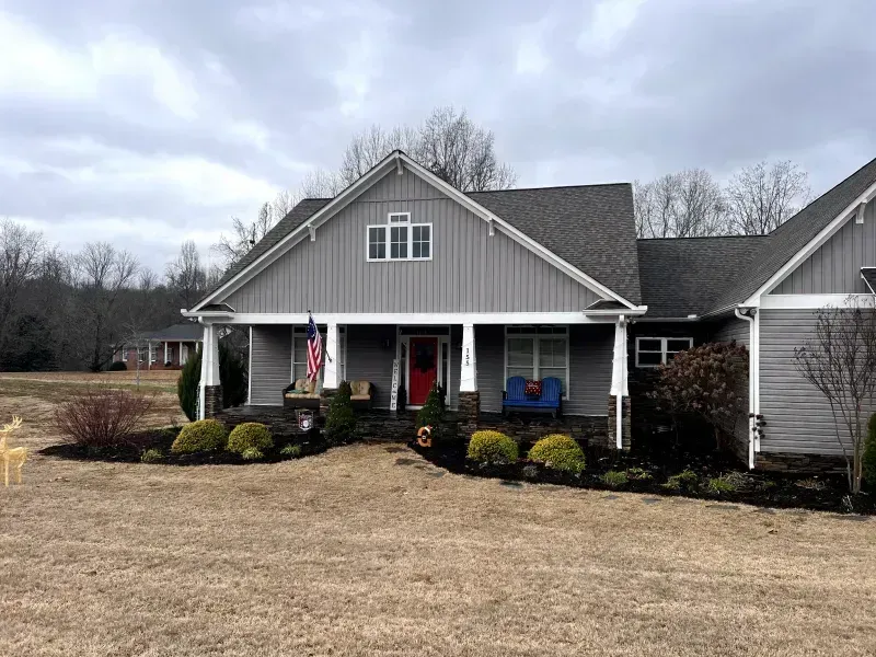Gray house with porch, red door, American flag, and dark gray roof. Cloudy day.