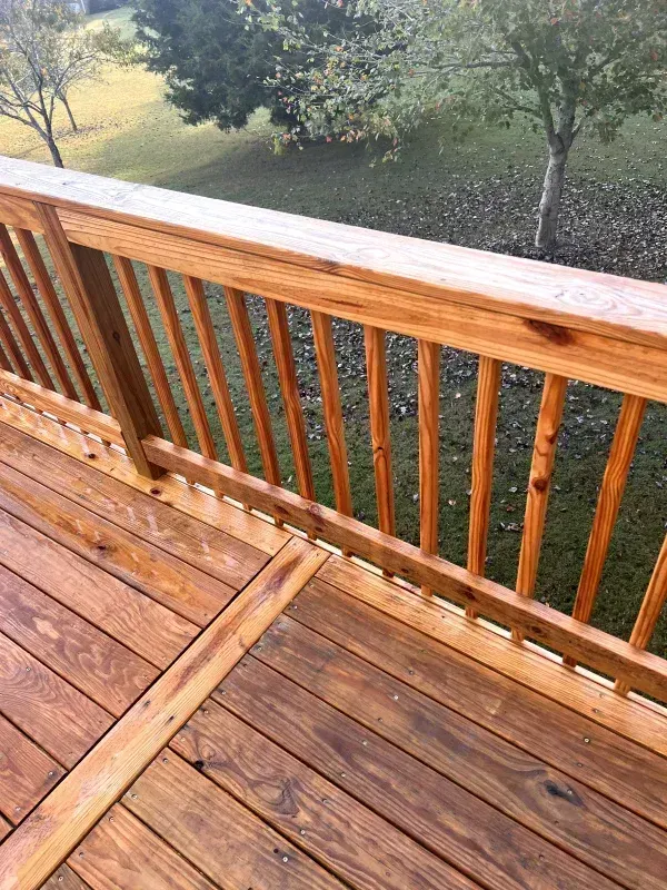 Wooden deck railing with vertical balusters, stained brown. Green grass and trees in the background.