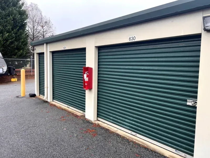Storage units with green metal roll-up doors, a fire extinguisher, and a numbered unit in an outdoor setting.