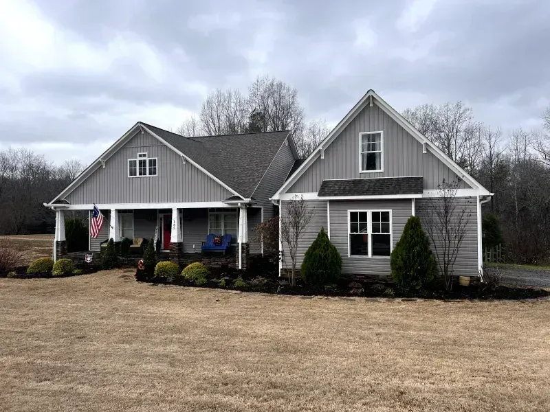 Gray house with white trim, porch, and a brown lawn under a cloudy sky.
