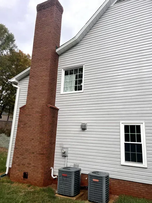 Brick chimney on a gray-sided house with two AC units and two windows.