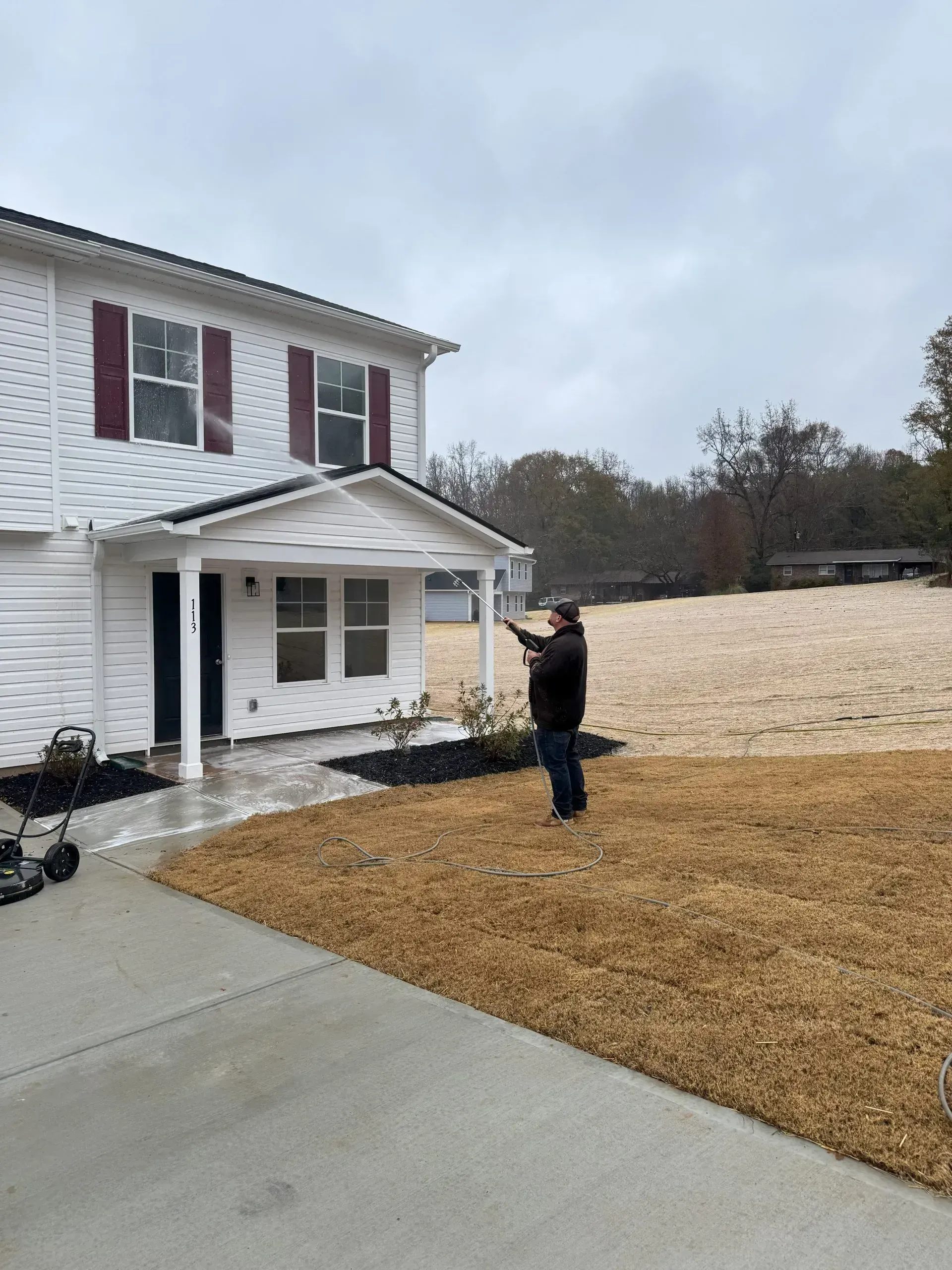 Man points toward a two-story white house with red shutters on a cloudy day, standing on brown grass.