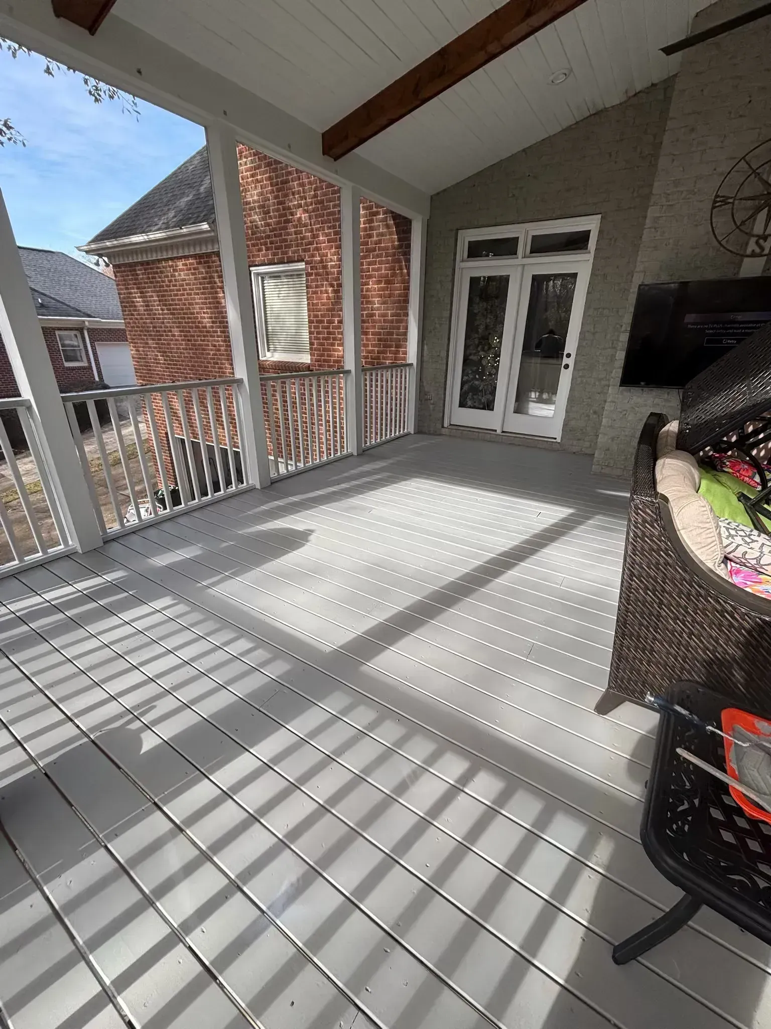 Covered porch with gray flooring and railing, view of brick building and double doors.