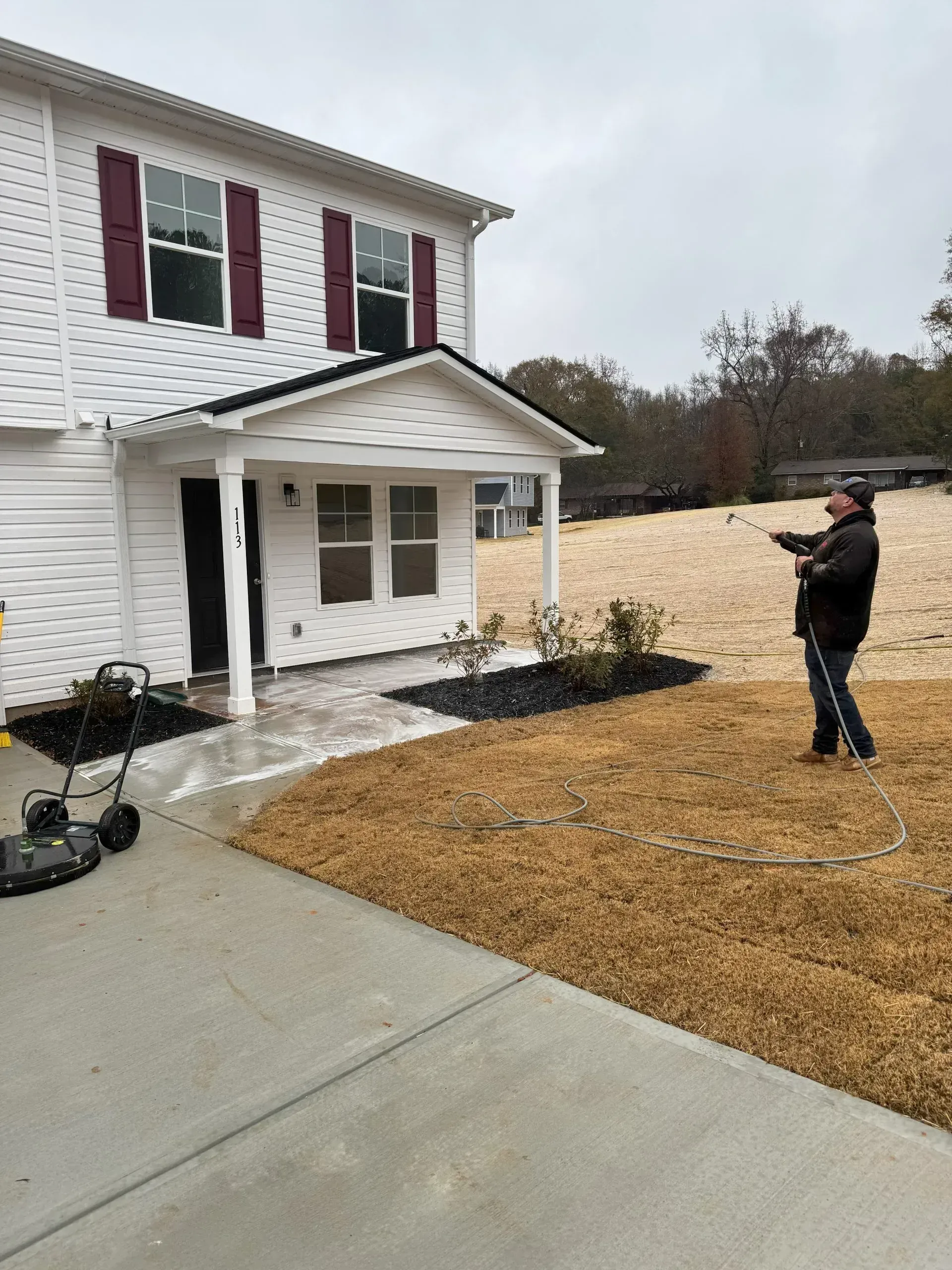 Man points toward house with white siding, burgundy shutters, and a covered porch; lawn mower nearby.