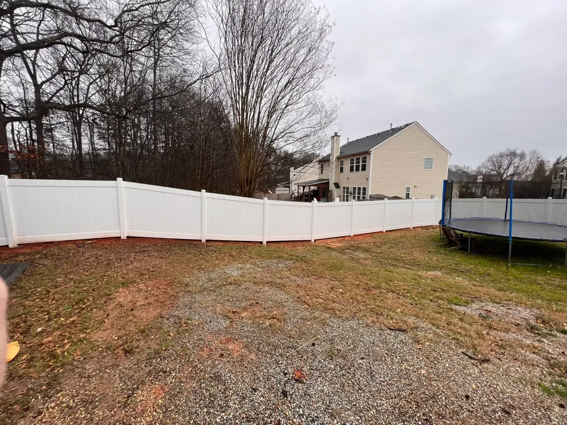 White fence in backyard with a house, a trampoline, and bare trees in the background.