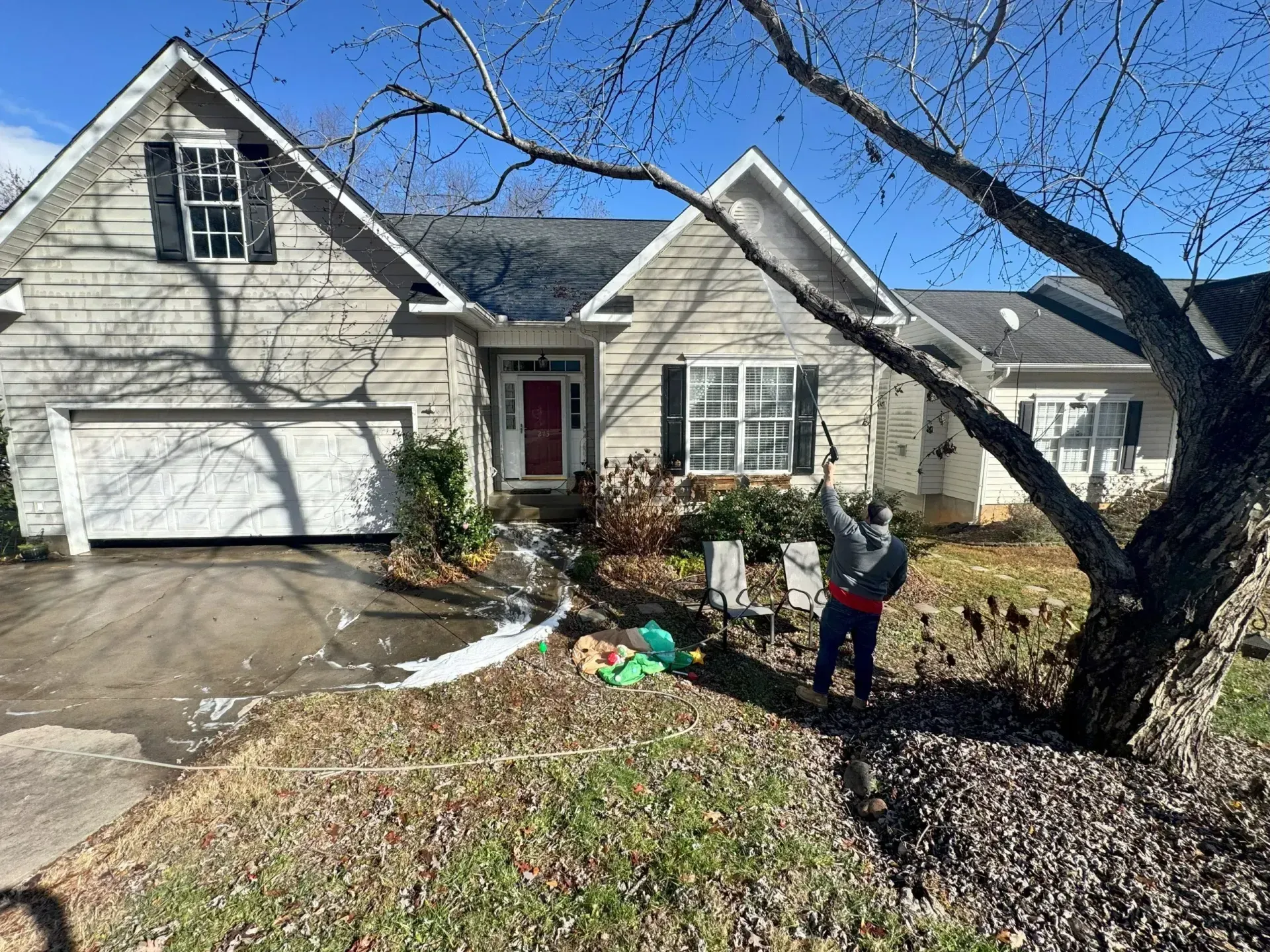 Person pressure washing a house with gray siding; driveway covered in foam. Sunny day.