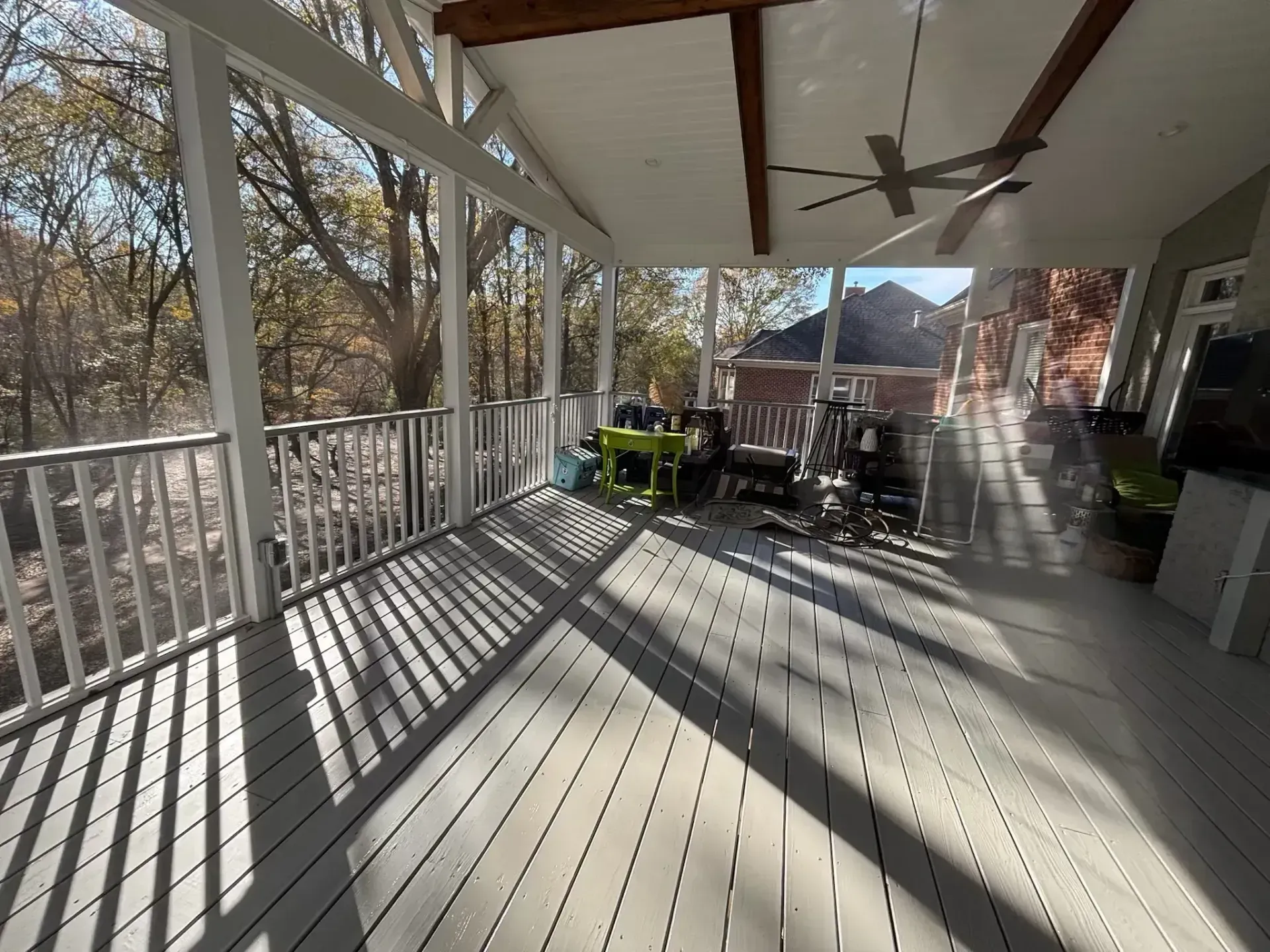 Screened-in porch with gray floorboards and white railings. Furniture sits in the background, with trees visible outside.
