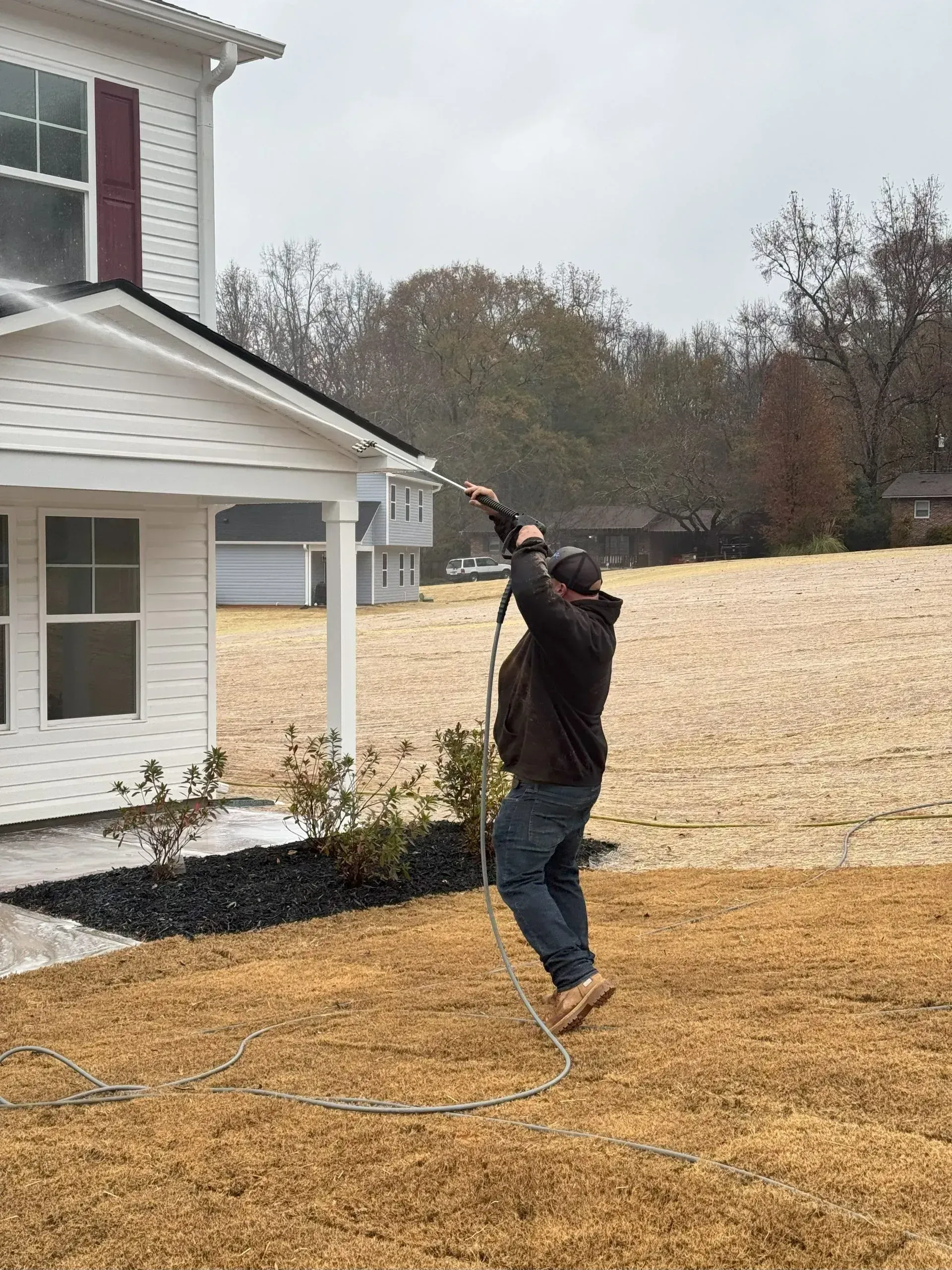 Person pressure washing a white house with brown roof and dry brown grass.