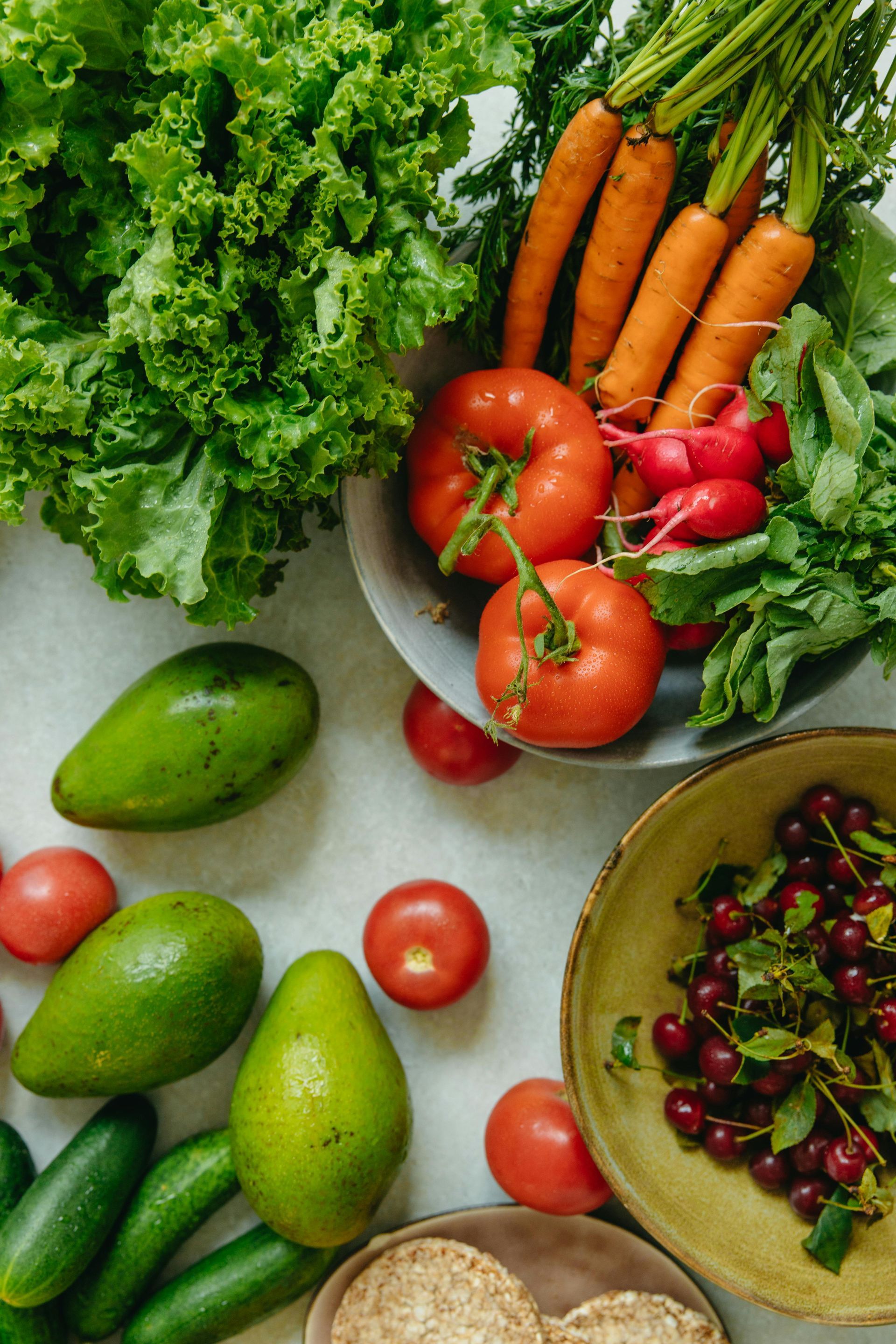 Variedad de verduras y frutas frescas sobre una mesa, incluyendo lechuga, zanahorias, aguacates, tomates y rábanos.