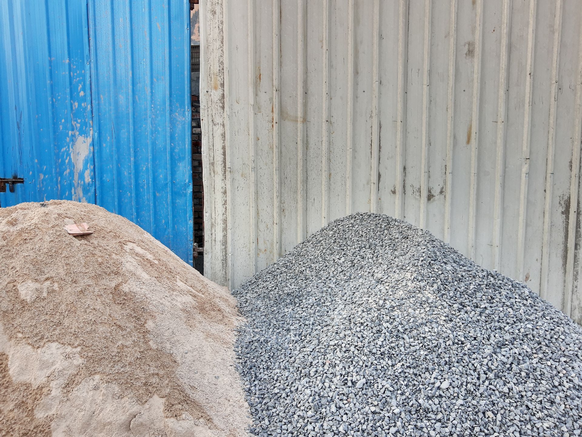 Pile of sand and pile of gravel stored side by side against a corrugated metal wall.