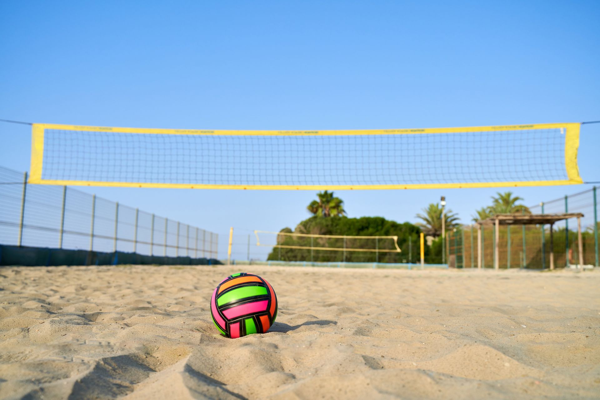 A volleyball is sitting on the sand near a volleyball net.
