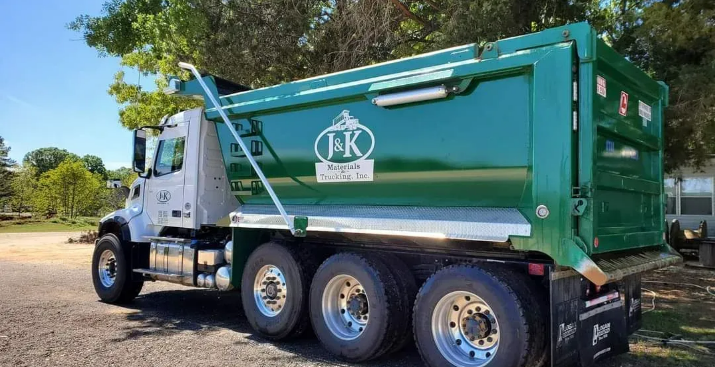 A truck from J & K Materials & Trucking is below a tree, at daylight.