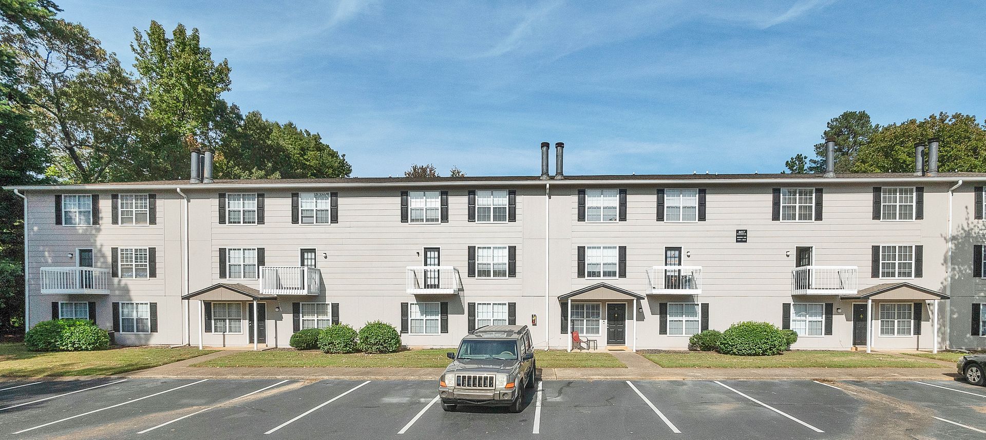 A three-story apartment building with beige siding, dark shutters, and small balconies, fronted by a parking lot.
