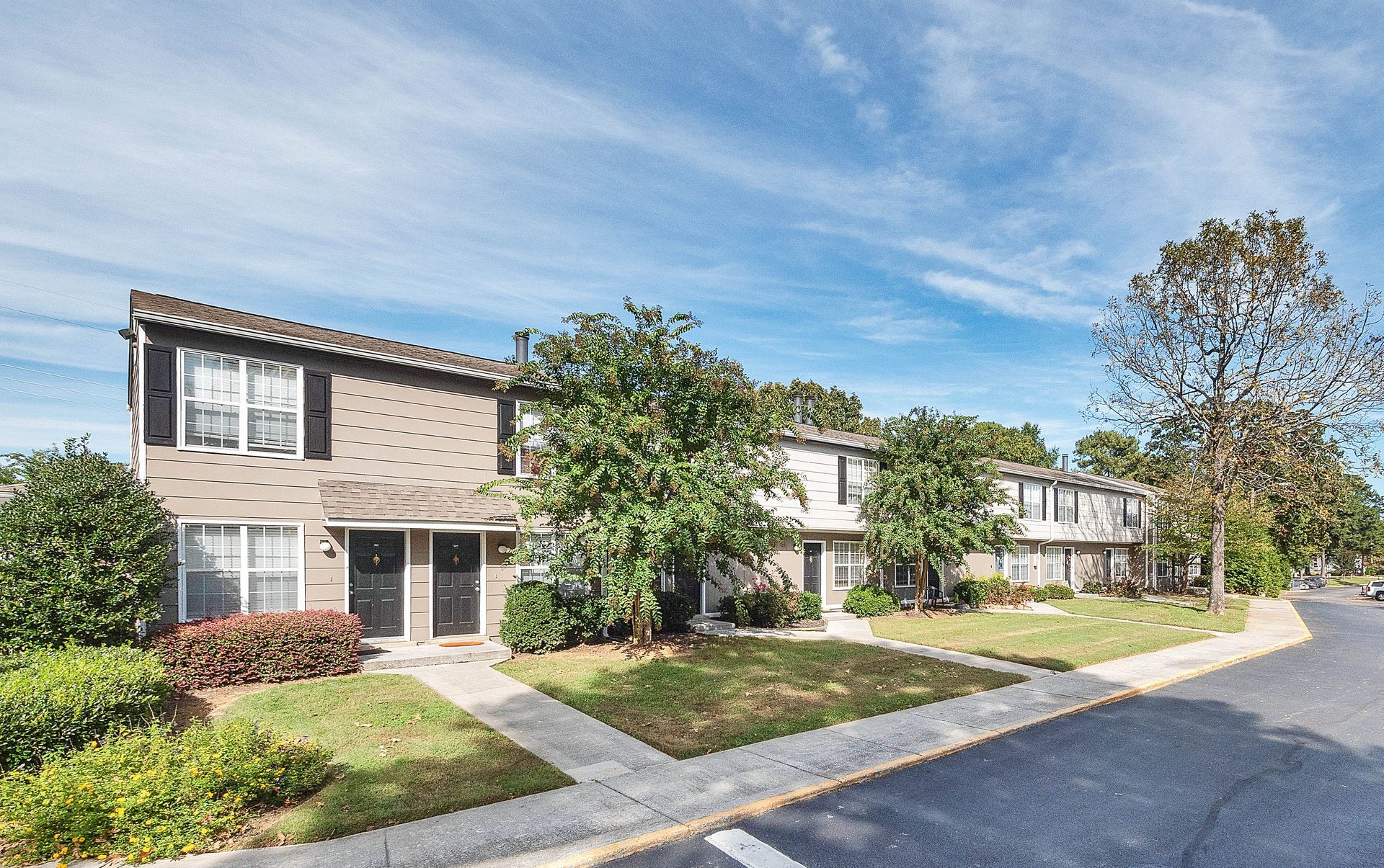 A multi-unit, two-story apartment building with tan siding, dark brown shutters, and green landscaping under a blue sky.