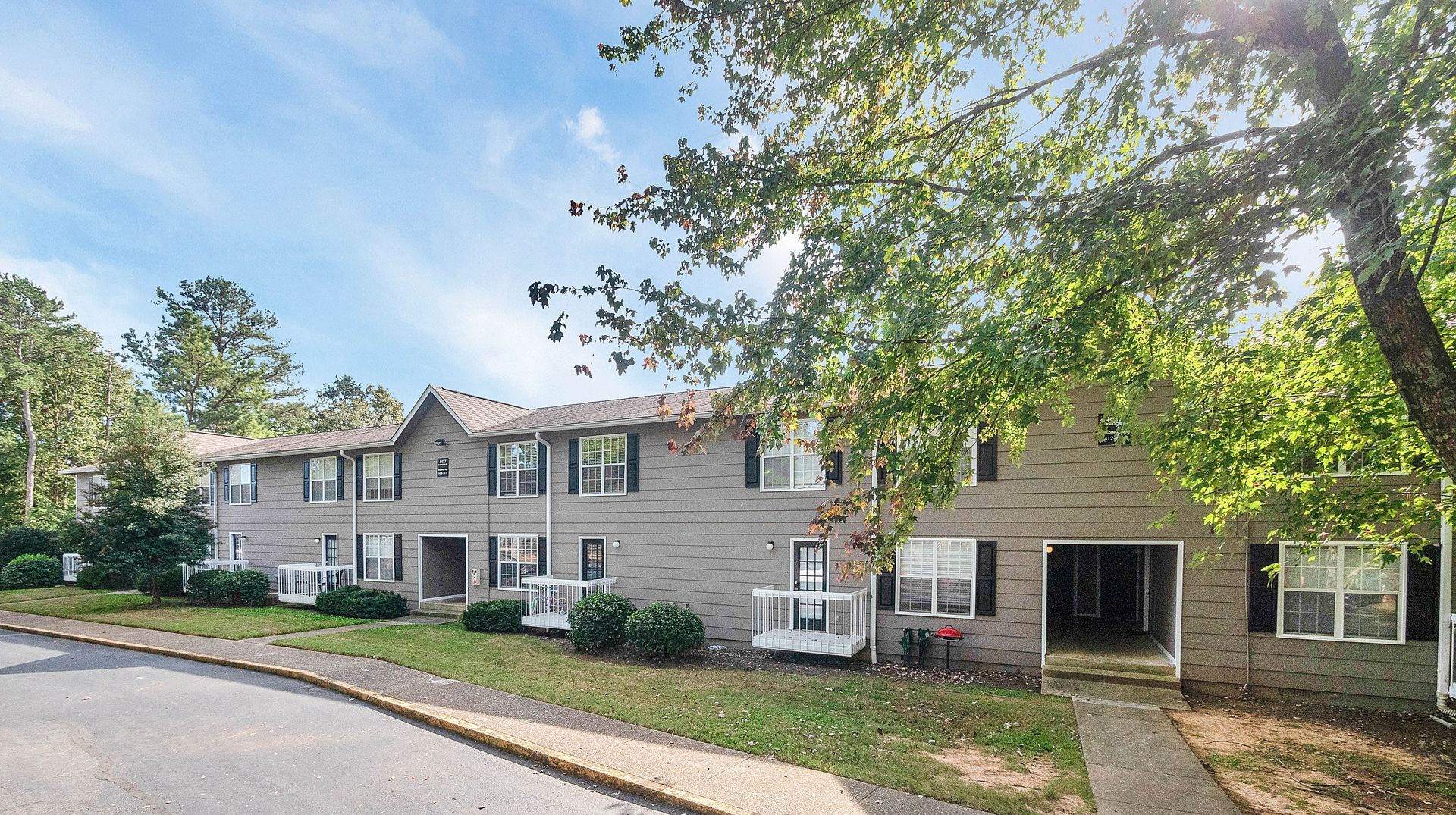 Two-story brick apartment building with white balconies and trim, surrounded by trees and a paved parking area.