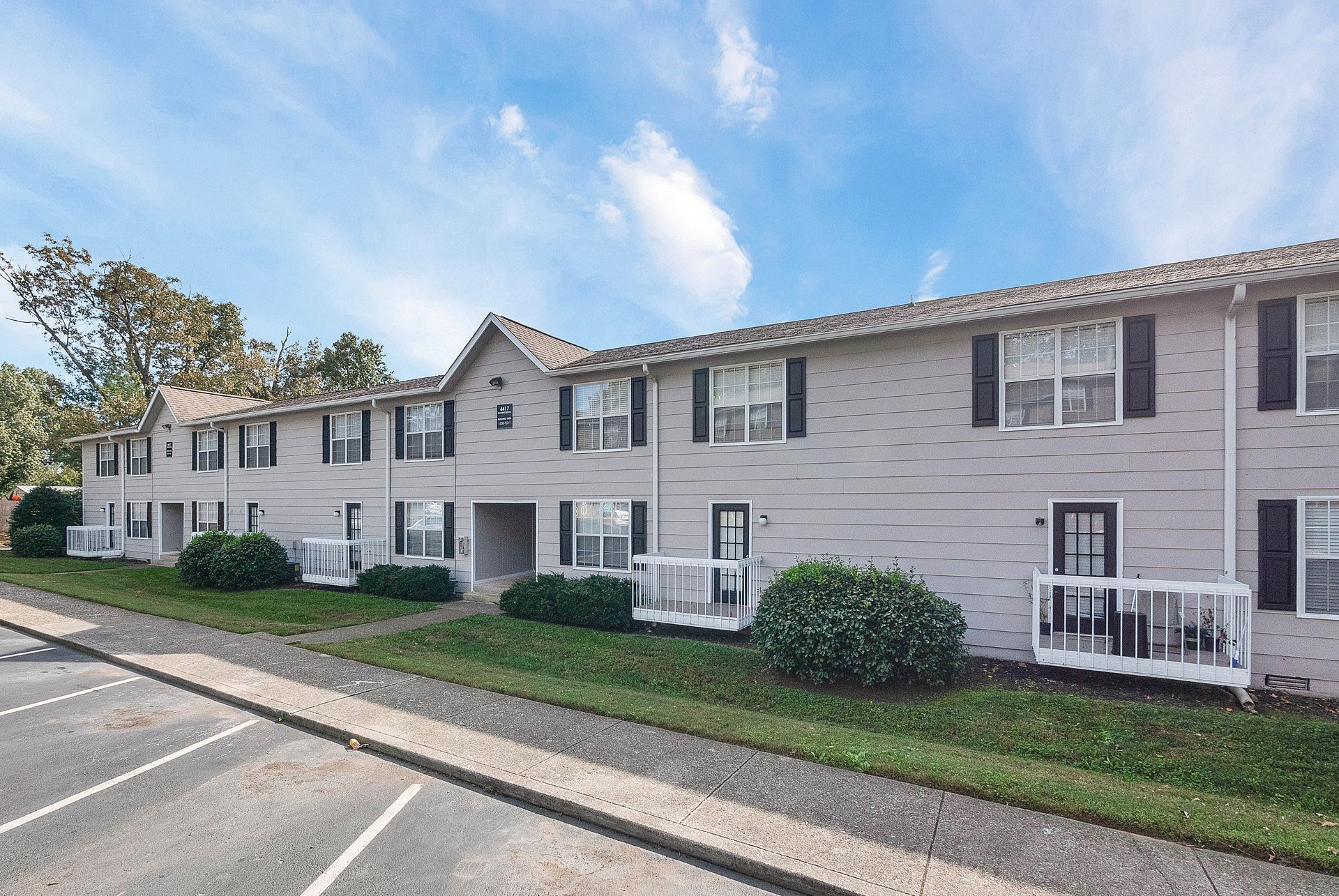 A two-story apartment complex with light-colored brick siding, white railings, and a parking lot in the foreground.