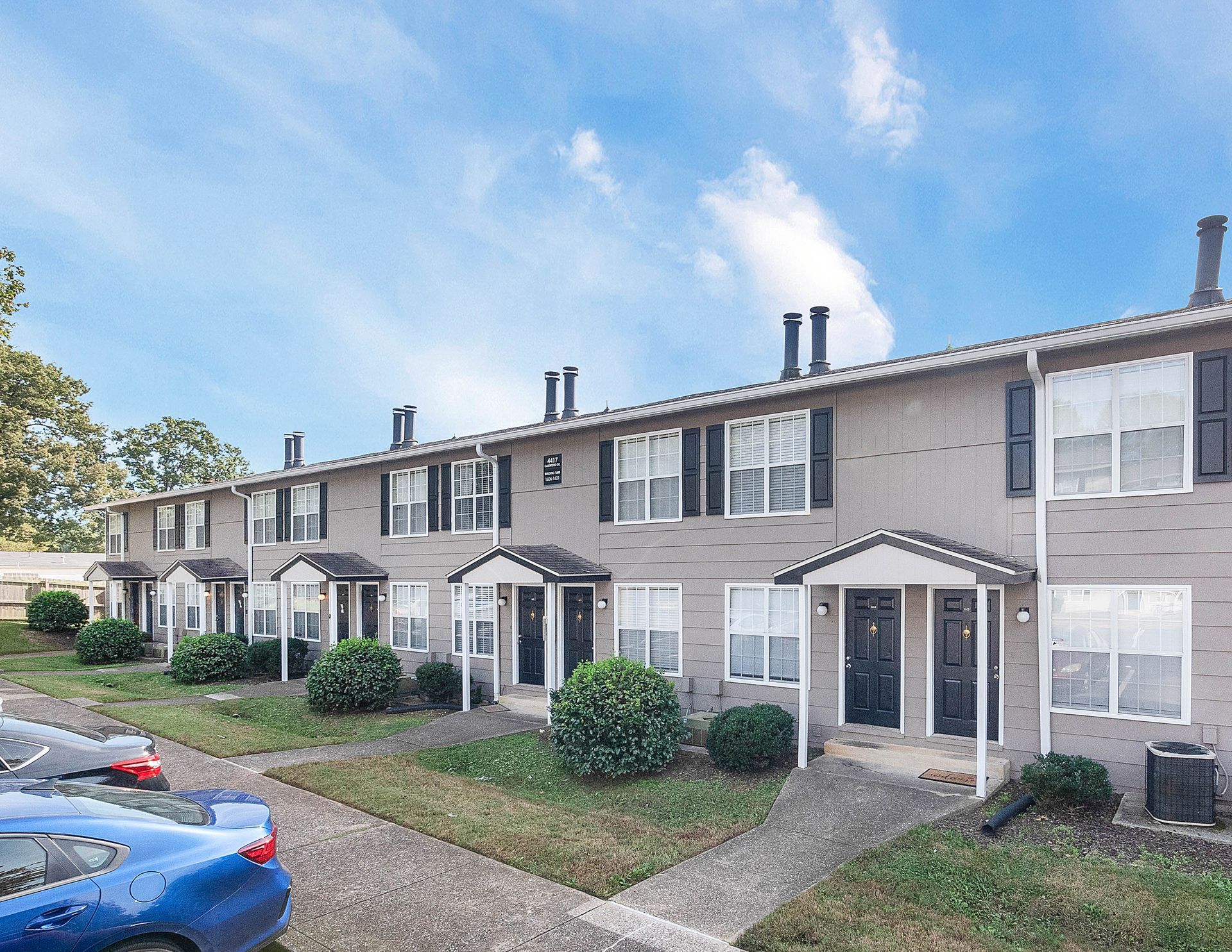 A row of tan, two-story apartment buildings with black shutters, dark doors, and small covered porches under a blue sky.