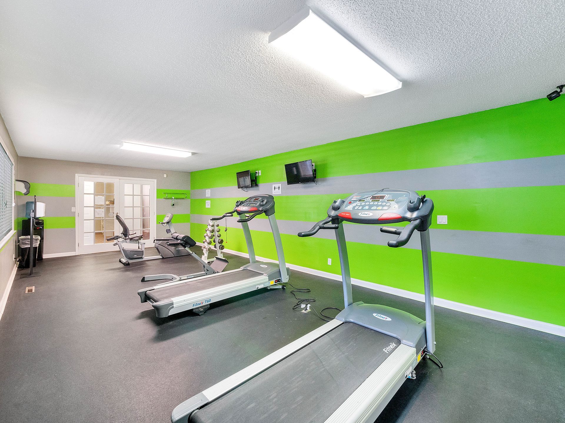 A gym interior with treadmills, a stationary bike, and grey and lime green striped walls under bright fluorescent lights.