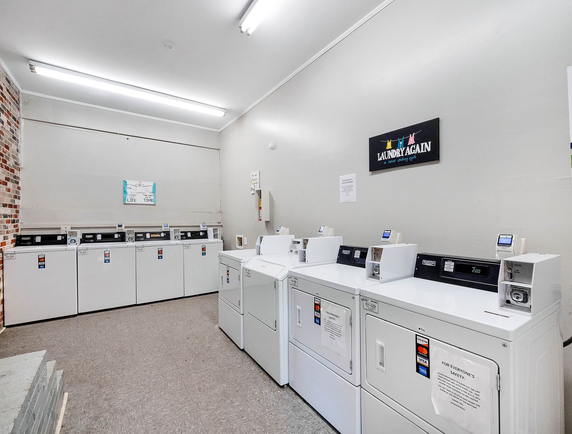 A laundry room with two rows of white coin-operated dryers against light grey walls under bright ceiling lights.