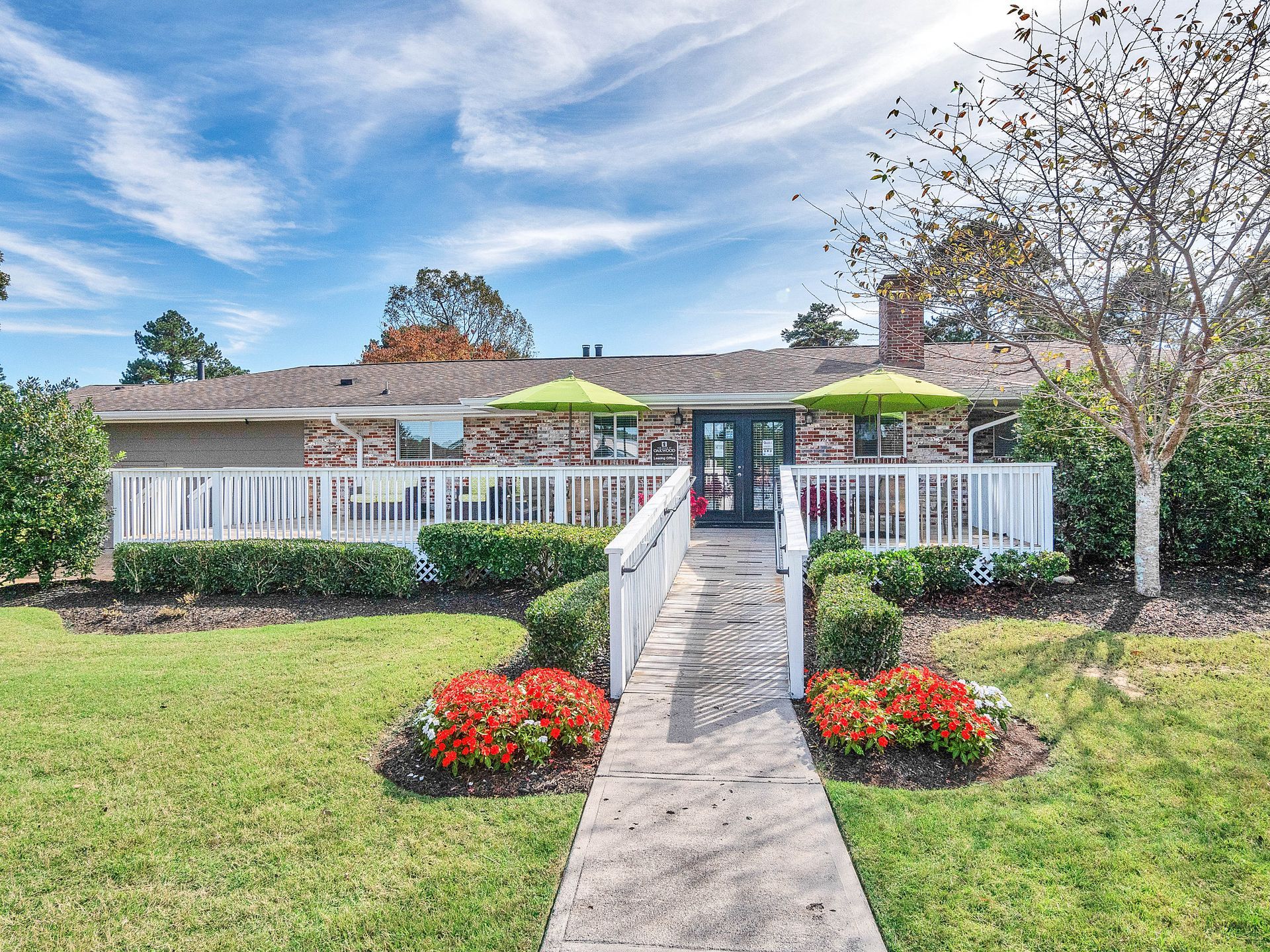 A brick home with a white deck, a concrete path leading to the entrance, and two green umbrellas on a sunny day.