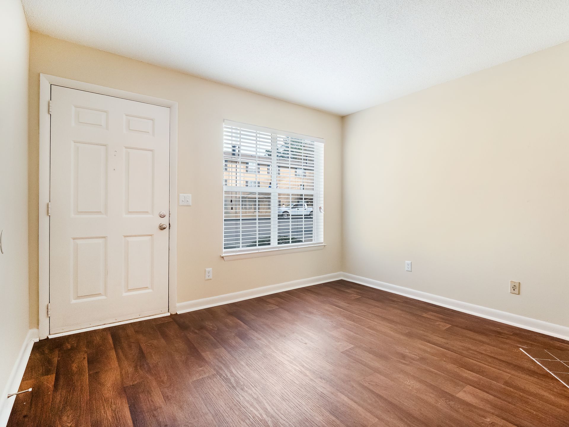 A bright room with light walls, wood-look flooring, a white front door, and a single window looking out at a building.