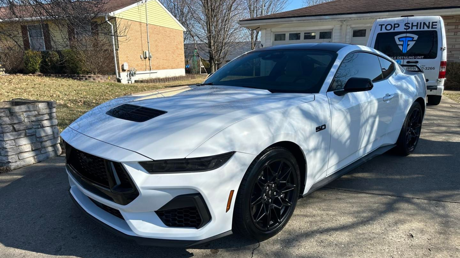 White Ford Mustang with black accents parked on a driveway in front of a house.