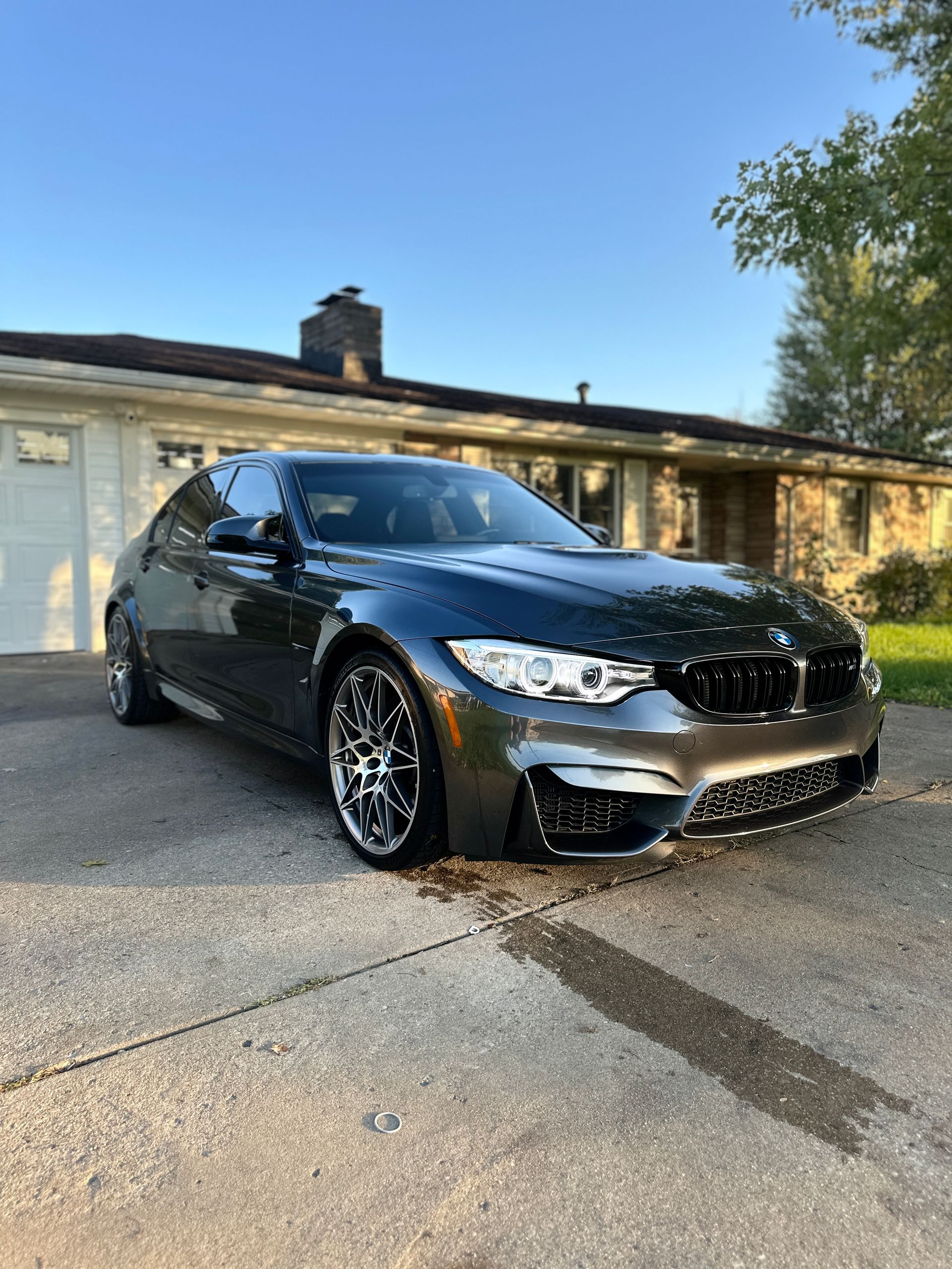 Dark gray BMW M3 sedan parked on a driveway in front of a house.