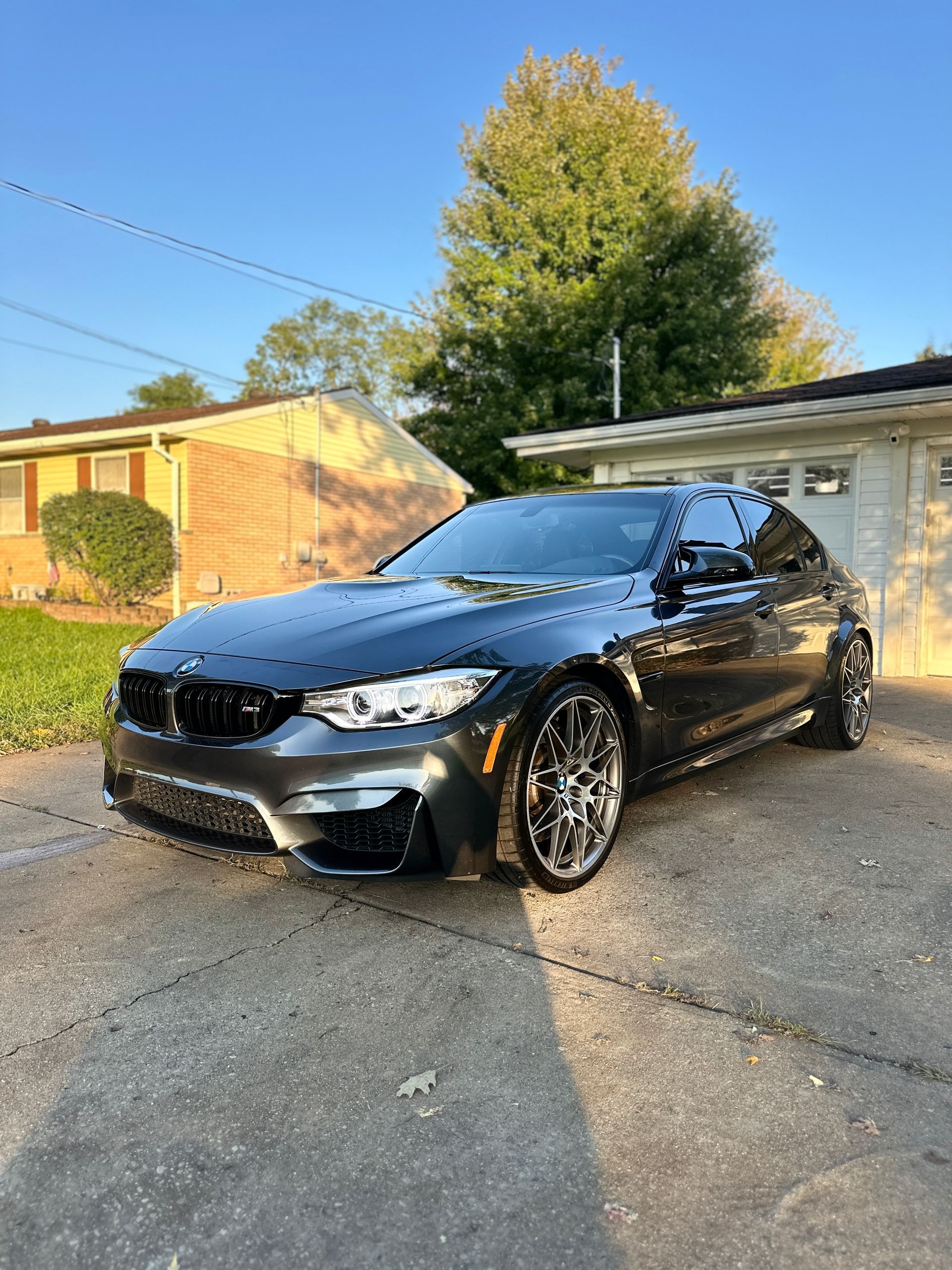 Dark gray BMW sports car parked on a concrete driveway in front of a house, bright sunny day.