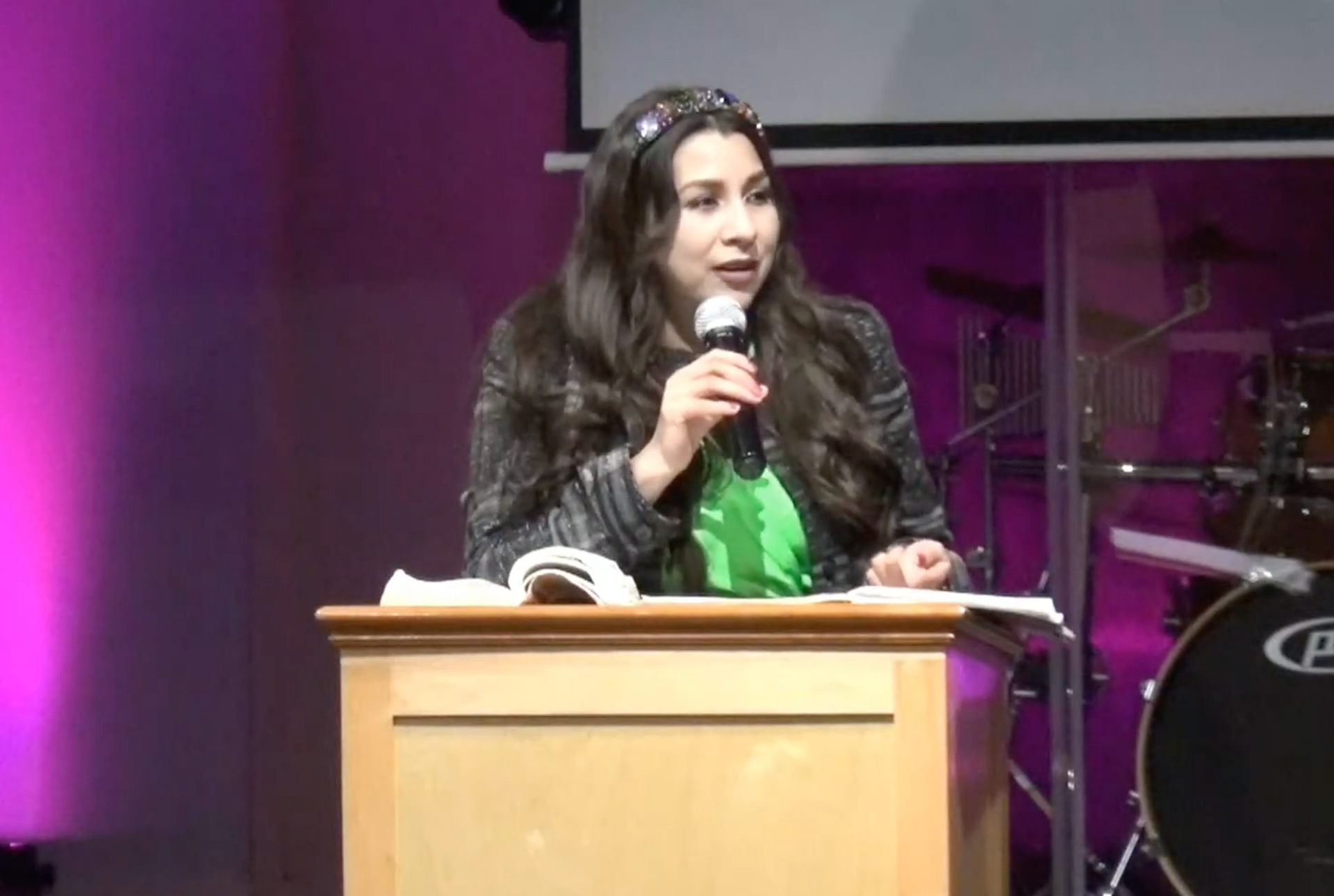 Una mujer habla por un micrófono mientras está de pie en un podio de madera frente a un escenario con iluminación púrpura y tambores.