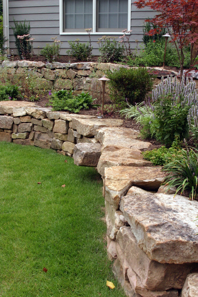 Stone retaining wall bordering a green lawn and garden in front of a house