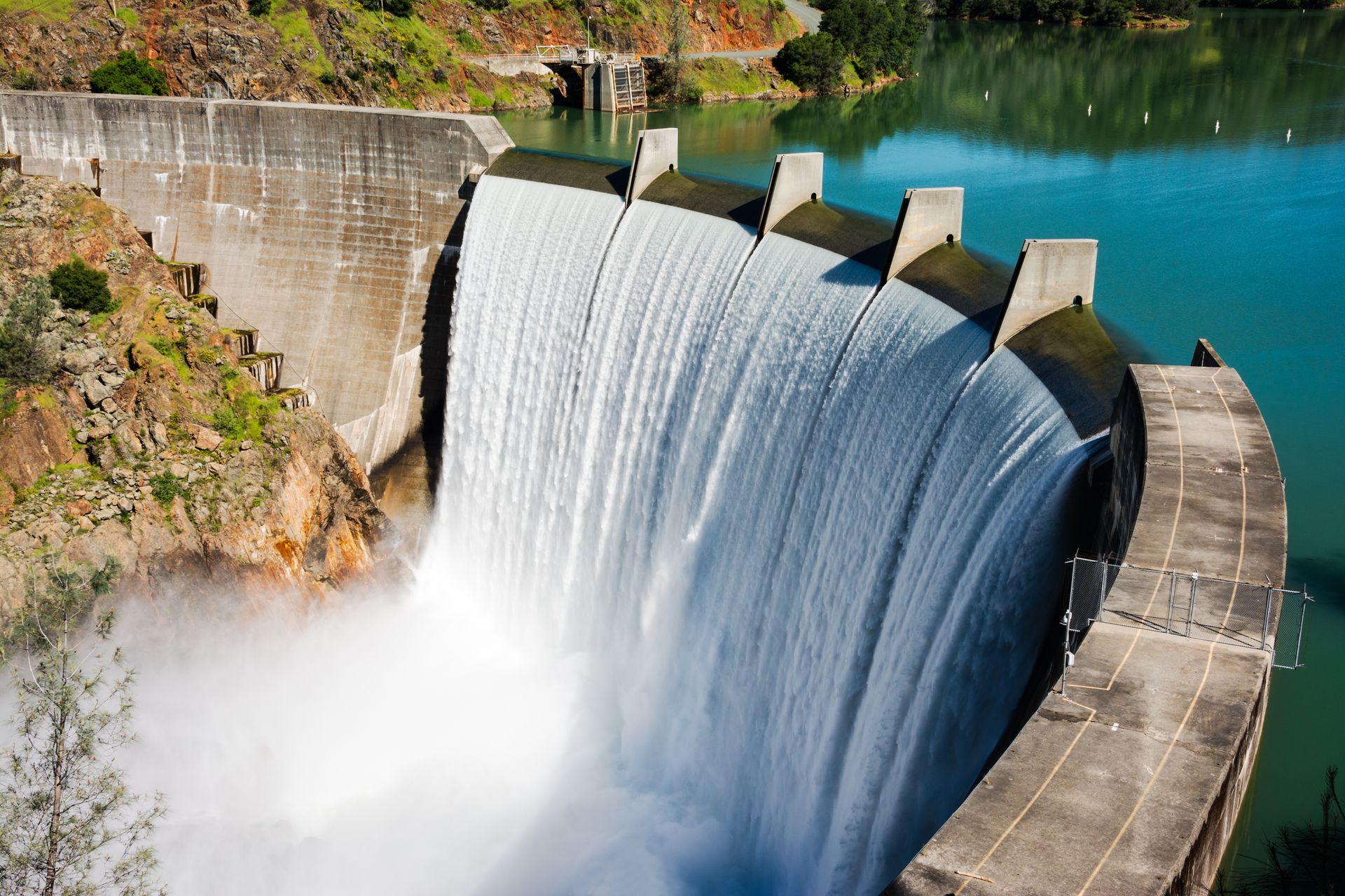 Dam with water cascading over its spillway into a turquoise lake.