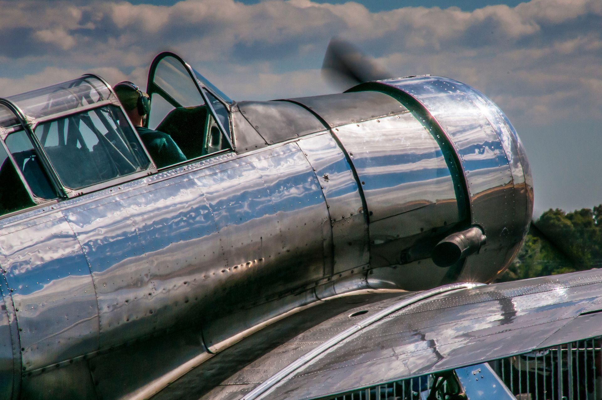 Shiny, silver airplane close-up with open cockpit; pilot visible; propeller spinning.