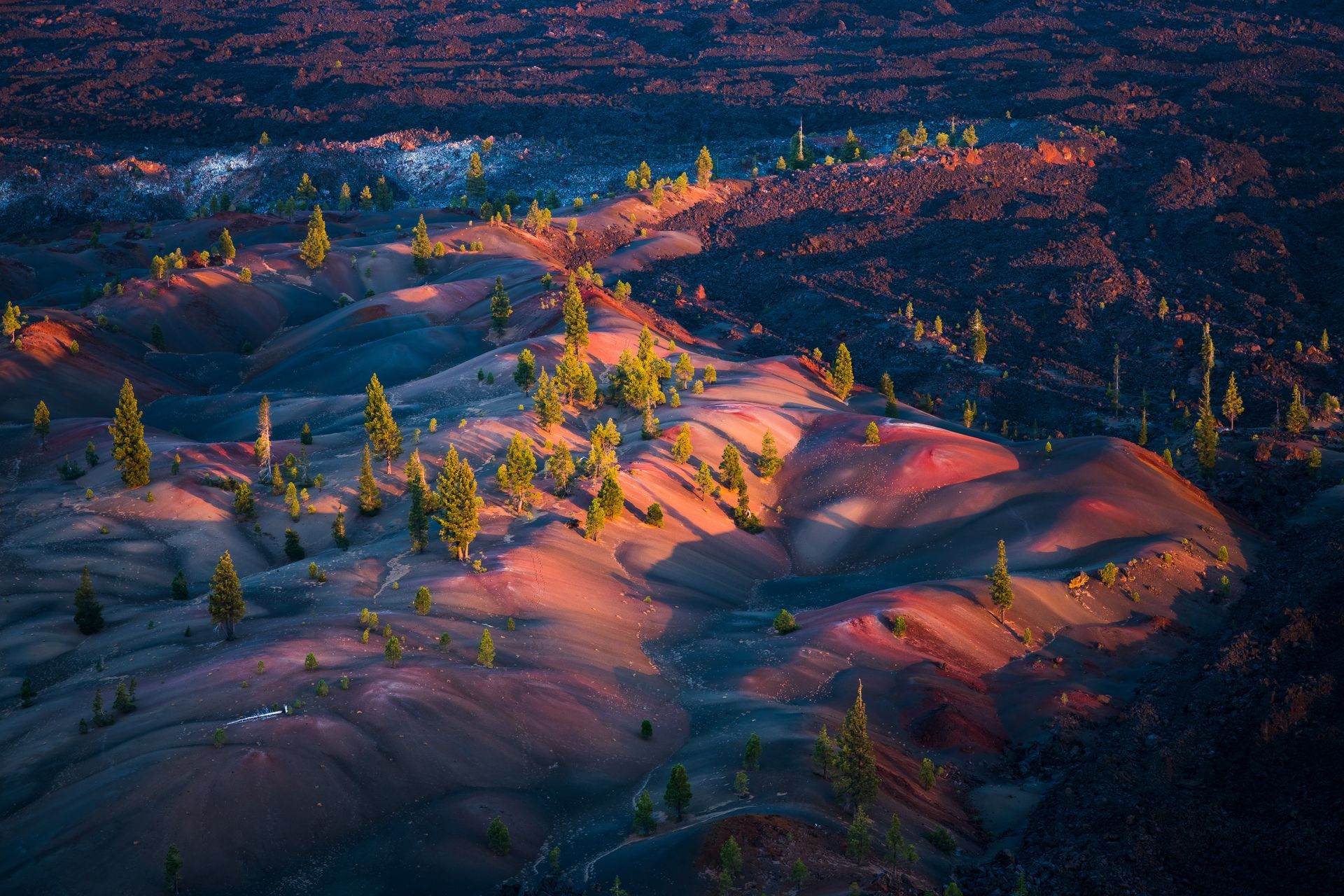 Sunset over a volcanic landscape with red and black rock formations and scattered green trees.