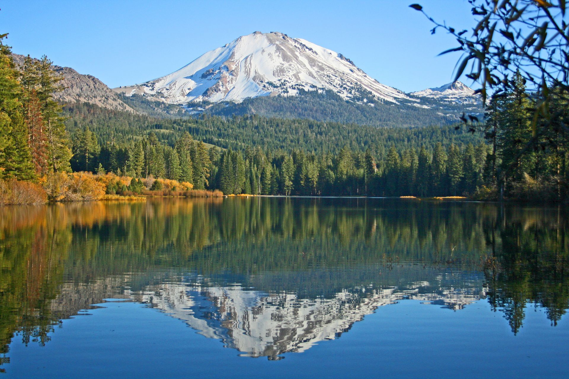 Snow-capped mountain reflecting in a calm lake, surrounded by evergreen trees and a clear blue sky.