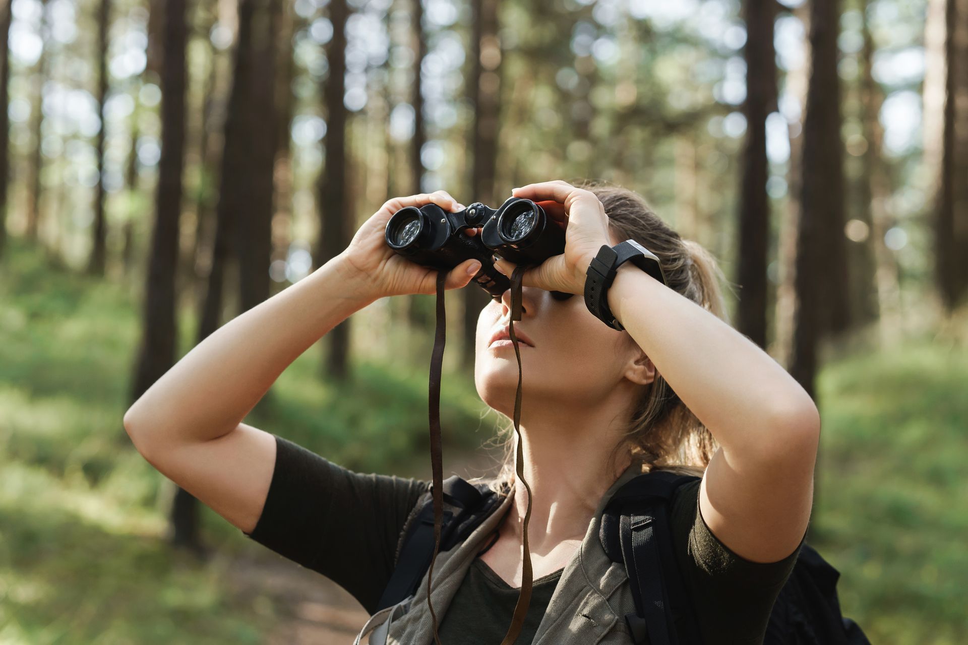 Woman looking through binoculars in a forest, with trees in the background.