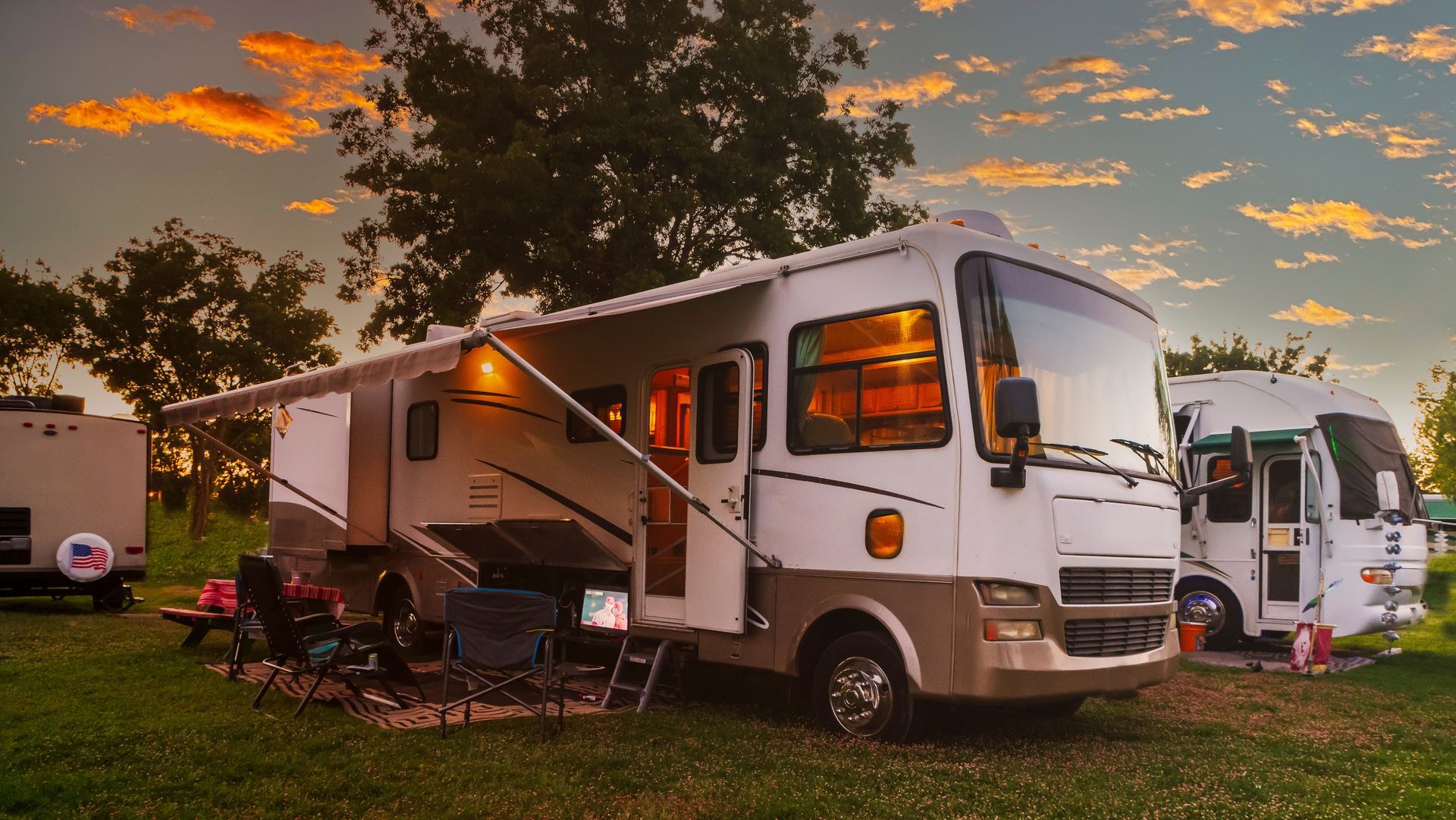 Two white RVs parked on grass at sunset with awnings out and a warm orange sky