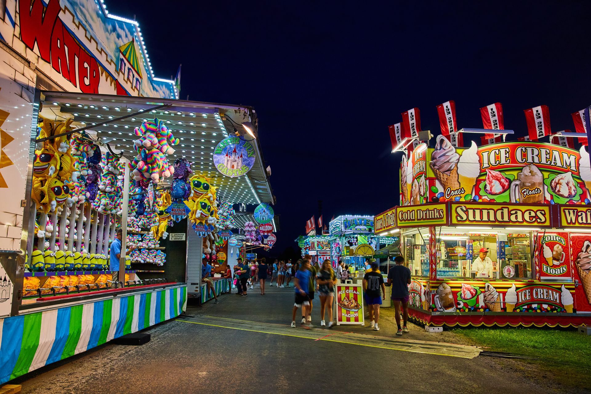 Night view of a brightly lit carnival midway featuring game booths and an ice cream stand with people walking between them.