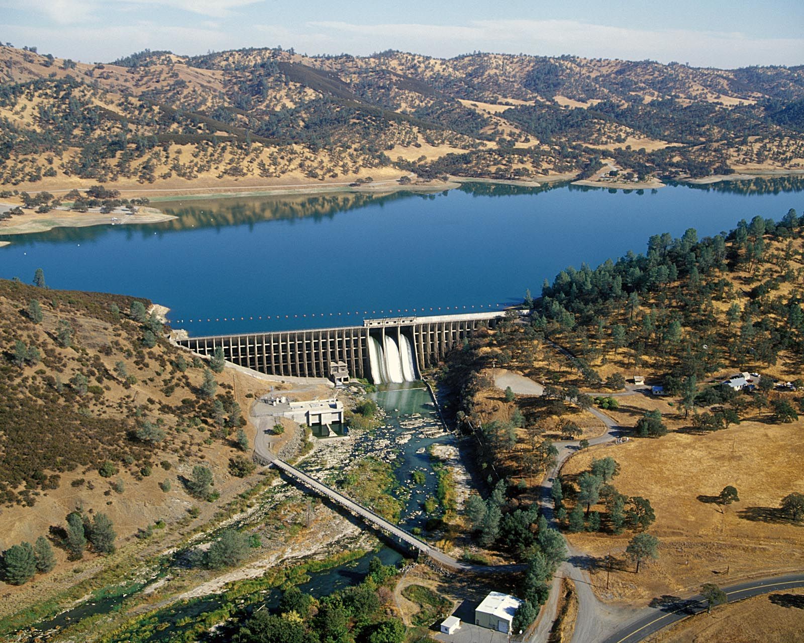 A dam with water flowing over its spillway into a river, surrounded by hills and a lake.