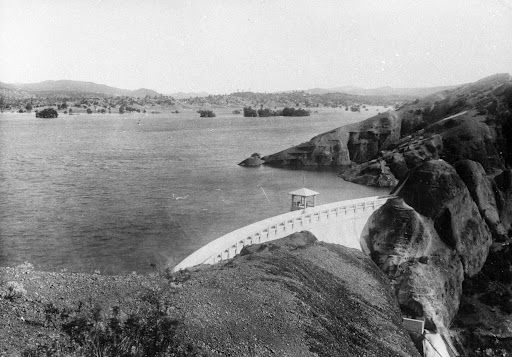 Black and white photo of a large dam. Water surrounds the dam with hills and trees in the background.