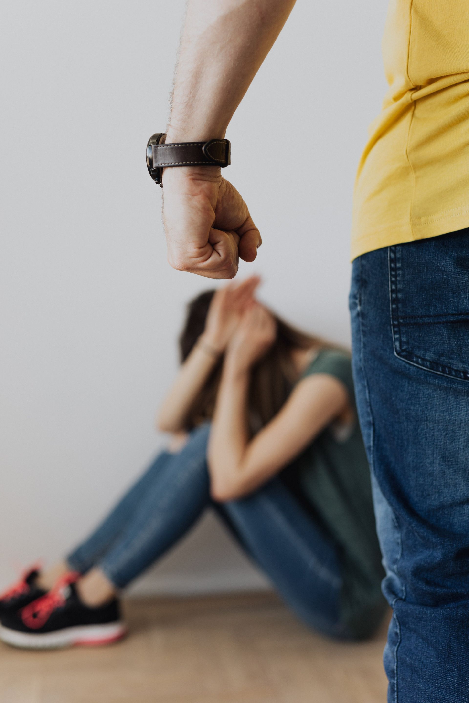 An image depicting a tense situation where a person in a yellow shirt and jeans has their clenched fist raised in a threatening gesture towards a woman sitting on the floor with her hands covering her face, appearing distressed. The focus is on the raised fist, conveying a powerful message about the intimidation and fear associated with domestic abuse.