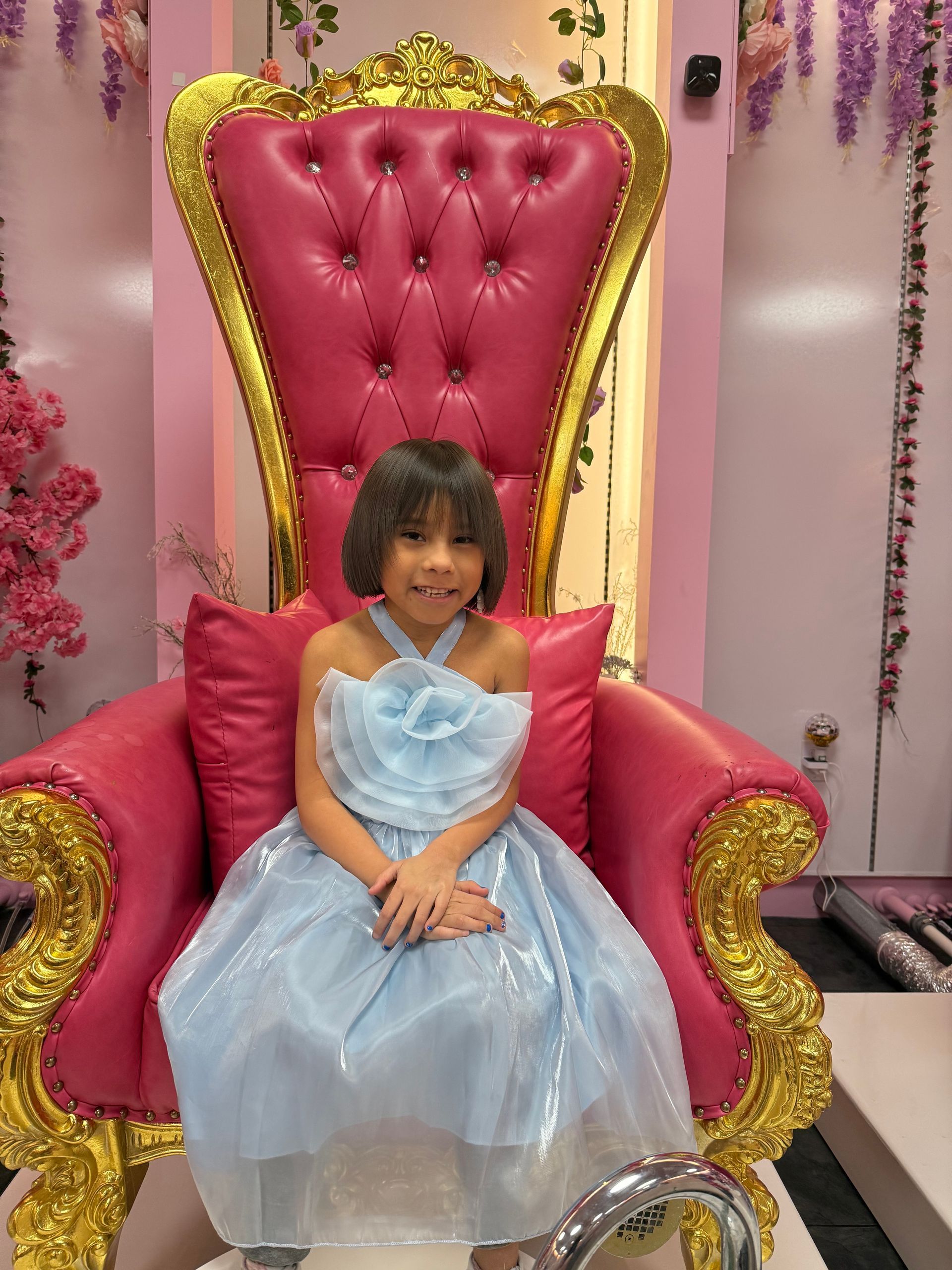 A little girl in a blue dress is sitting in a pink chair.