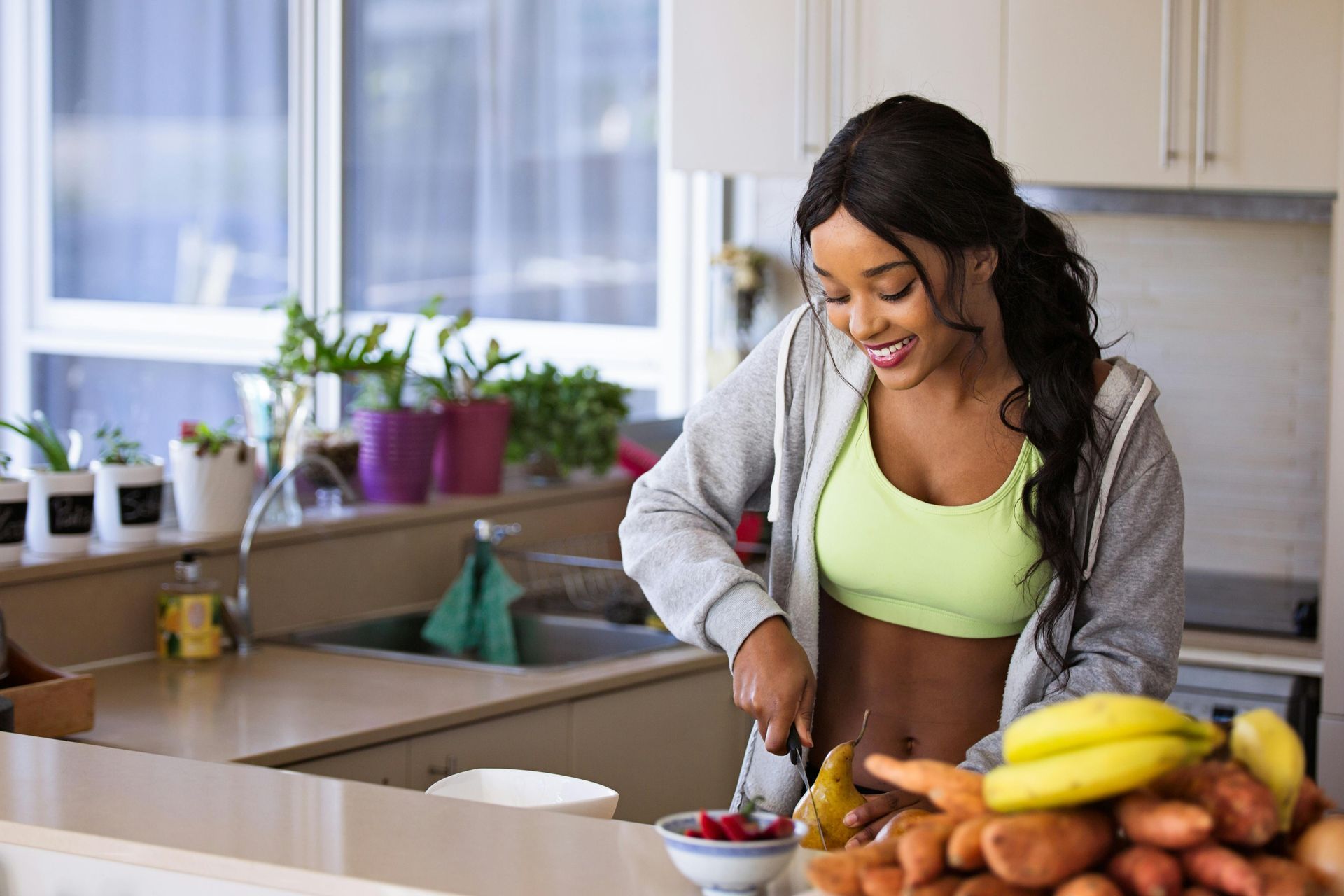 Woman smiling while chopping food in a kitchen. She's wearing a grey jacket, sports bra, and has a bowl of fruit nearby.