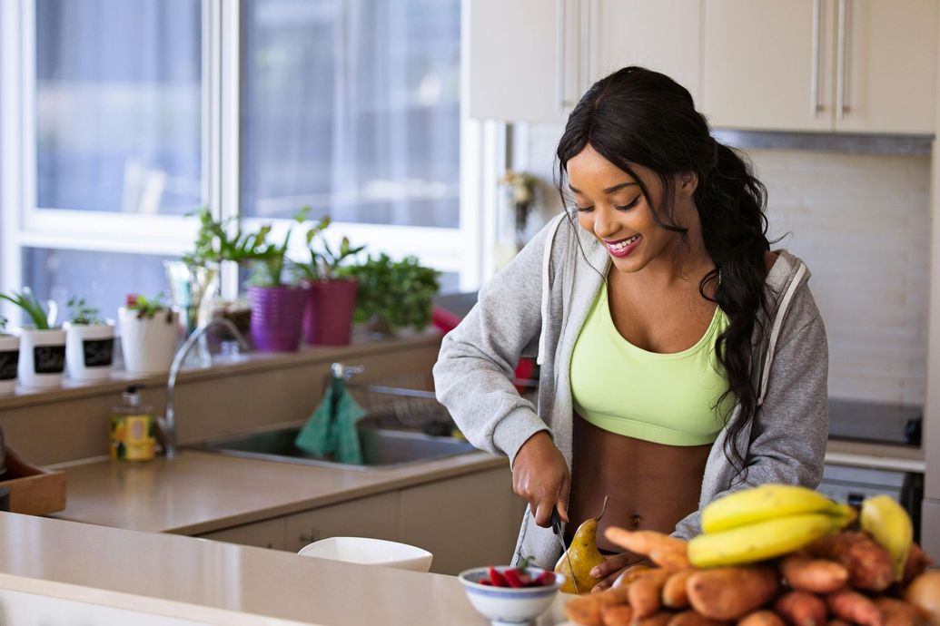 Woman smiling while chopping food in a kitchen. She's wearing a grey jacket, sports bra, and has a bowl of fruit nearby.