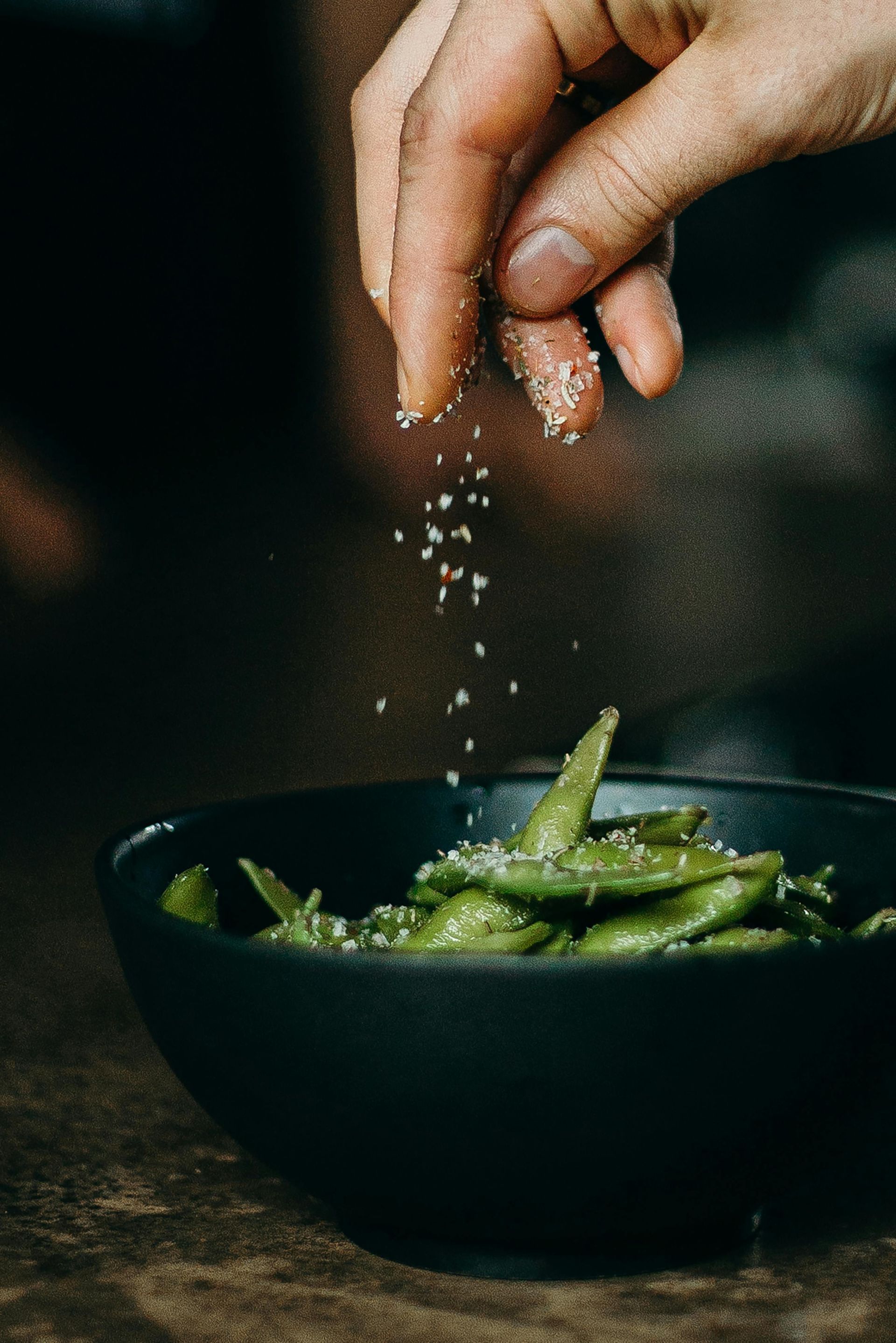 Hand sprinkling salt onto a black bowl of green edamame.