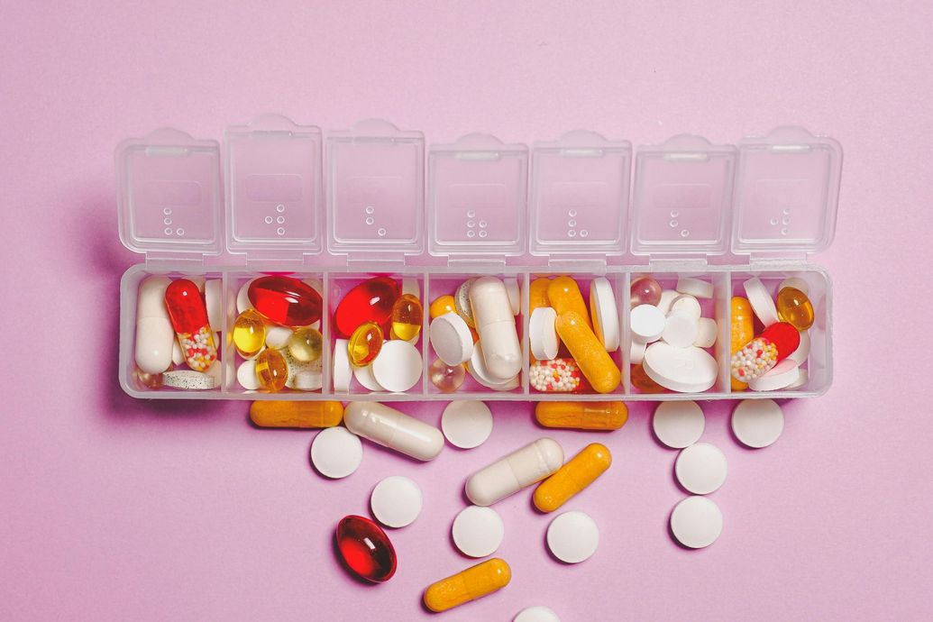 Pill organizer filled with colorful pills and capsules on a pink surface. Some pills are scattered.