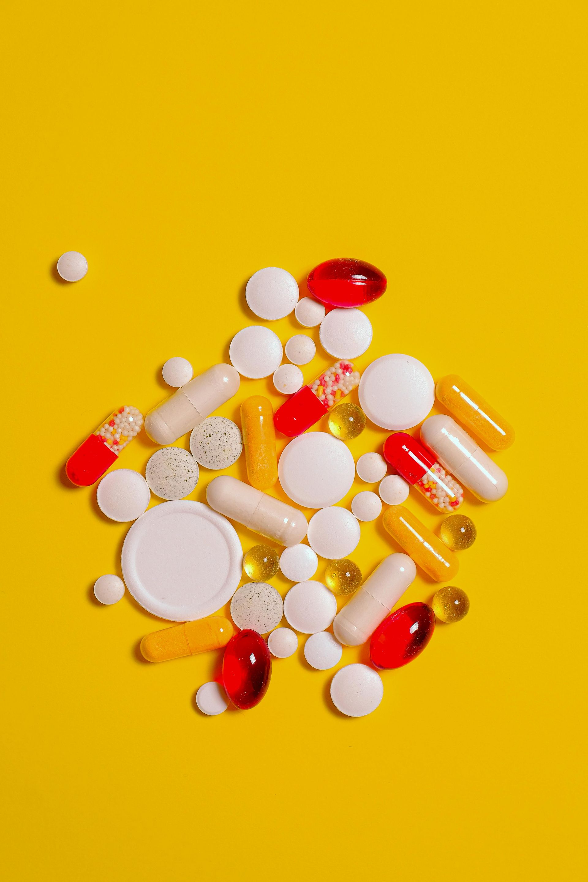 Pills and capsules scattered on a yellow surface, various shapes, sizes, and colors.