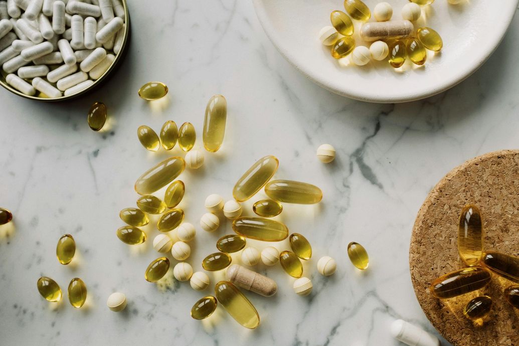 Assorted pills and capsules, including yellow softgels, scattered on a white marble surface, some in dishes.