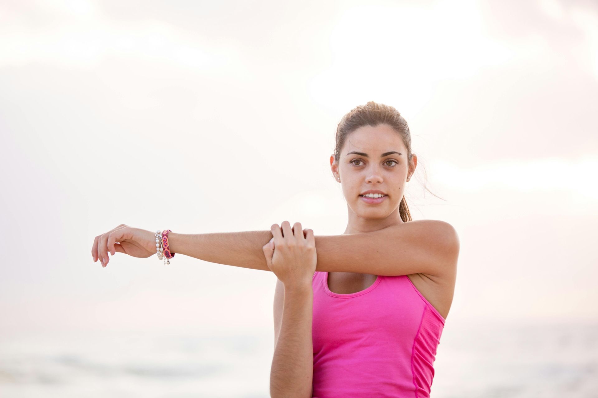 Woman in pink top stretching her arm, outdoors near water.