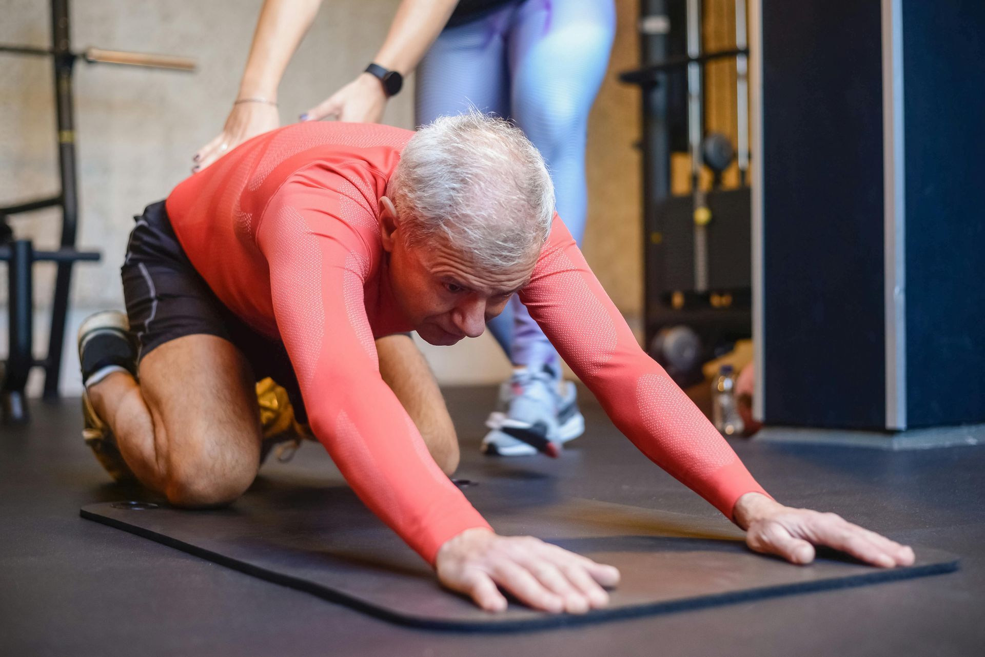 Man kneeling on mat, stretching with assistance in gym.