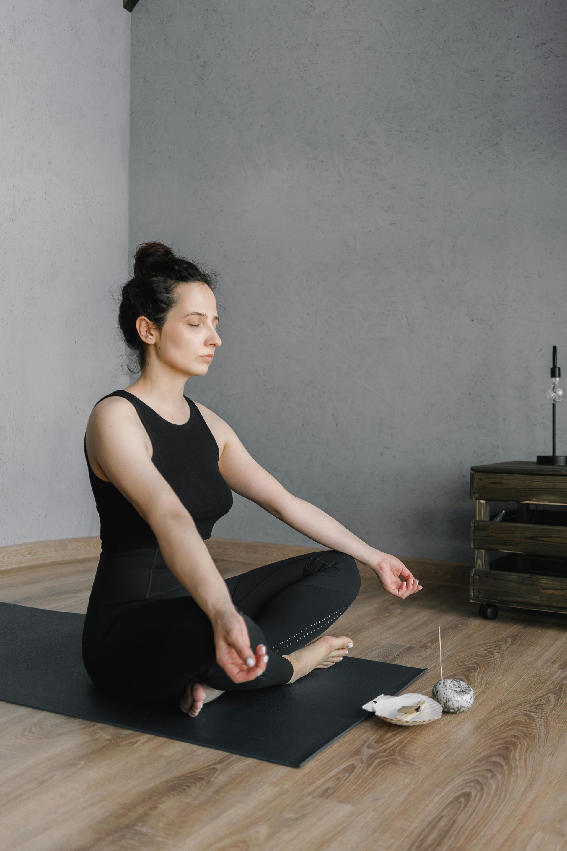 Woman in black yoga outfit meditates on a mat in a room with grey walls and wooden floor.
